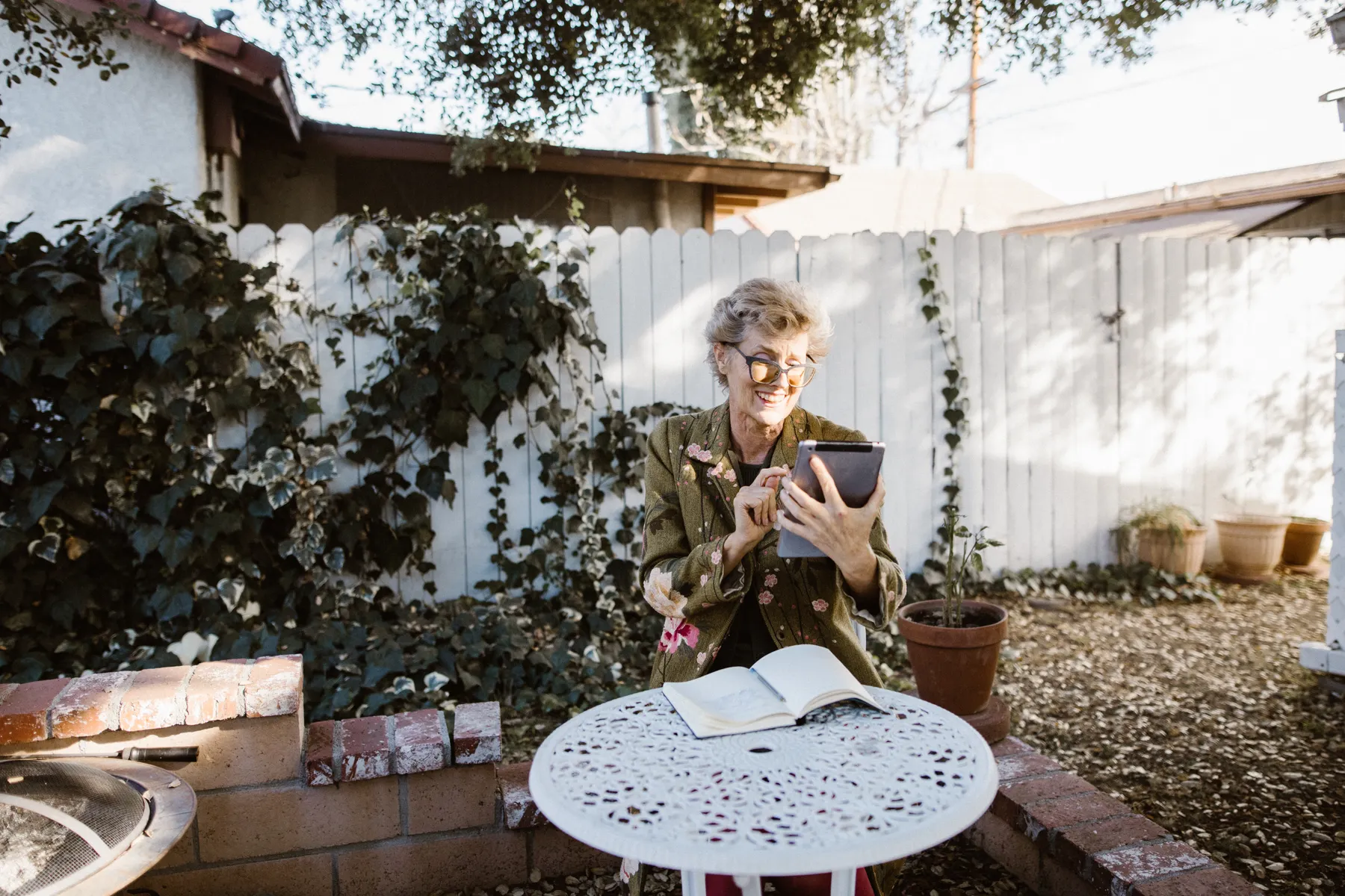 An older woman smiling while using a tablet in a sunny garden